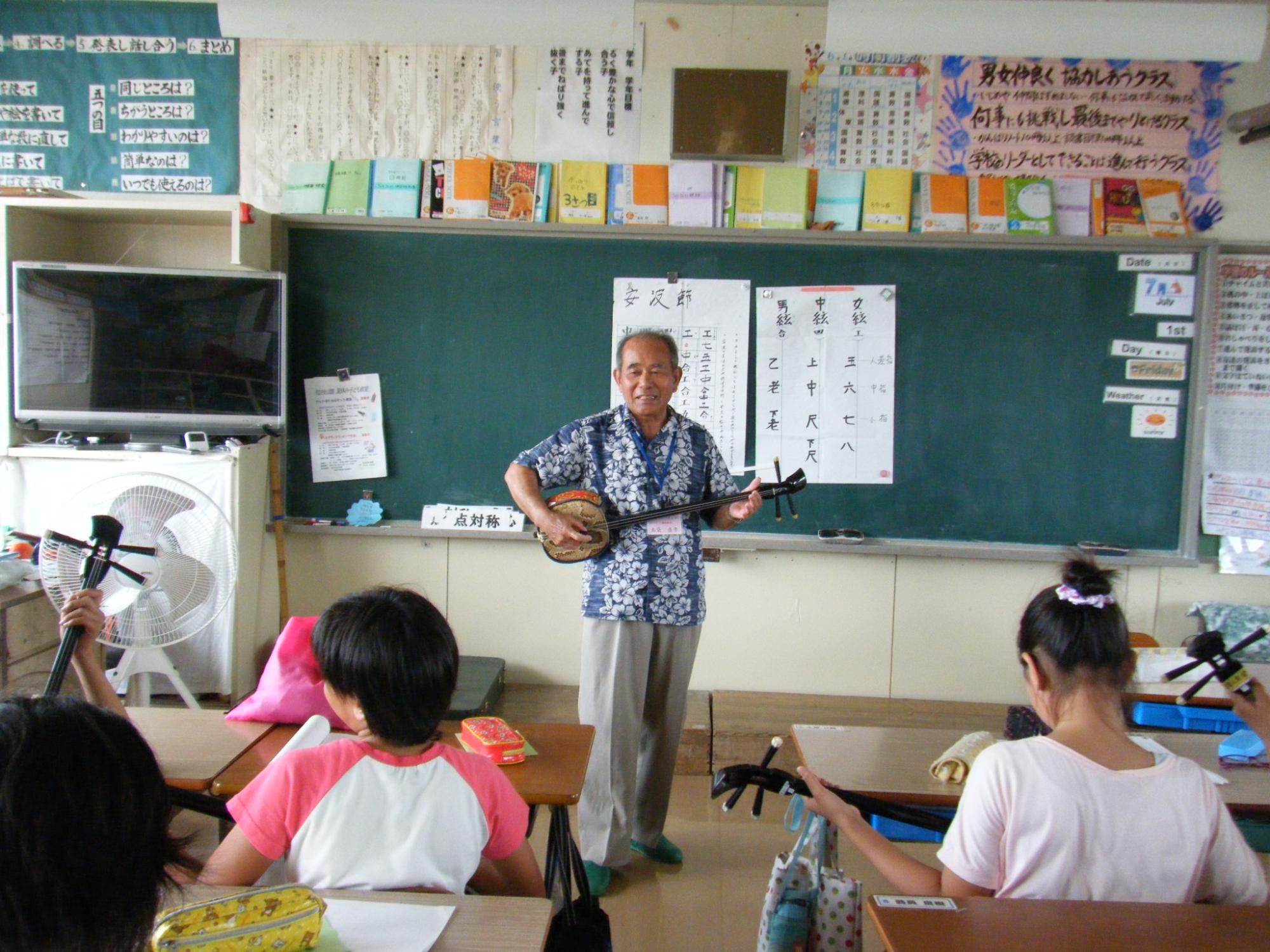 学校の黒板の前で数人の子供を前に立って三味線を弾いている老人の写真