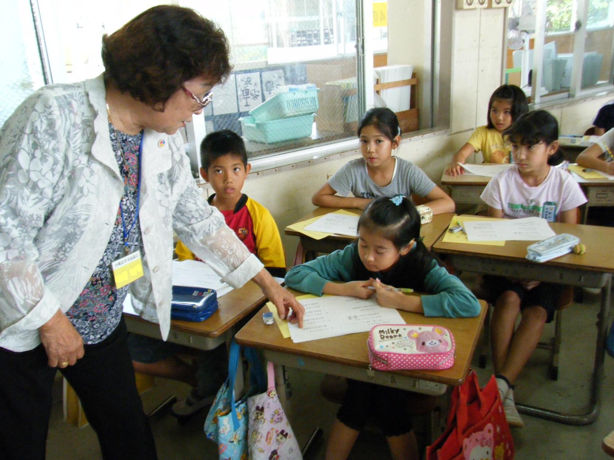 学校の教室で机にプリントを広げて座る子供にプリントに指をさして話をしている老人の写真