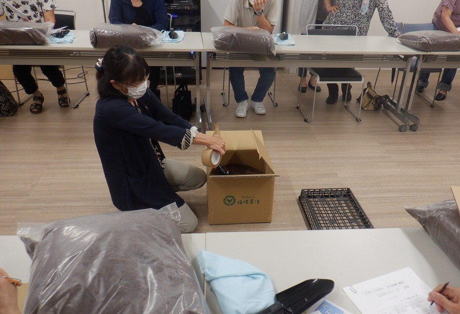 ダンボールに入っている堆肥を抱えた女性がこの字型に並べた机に座って講習を受けている人々に中身を見せている写真