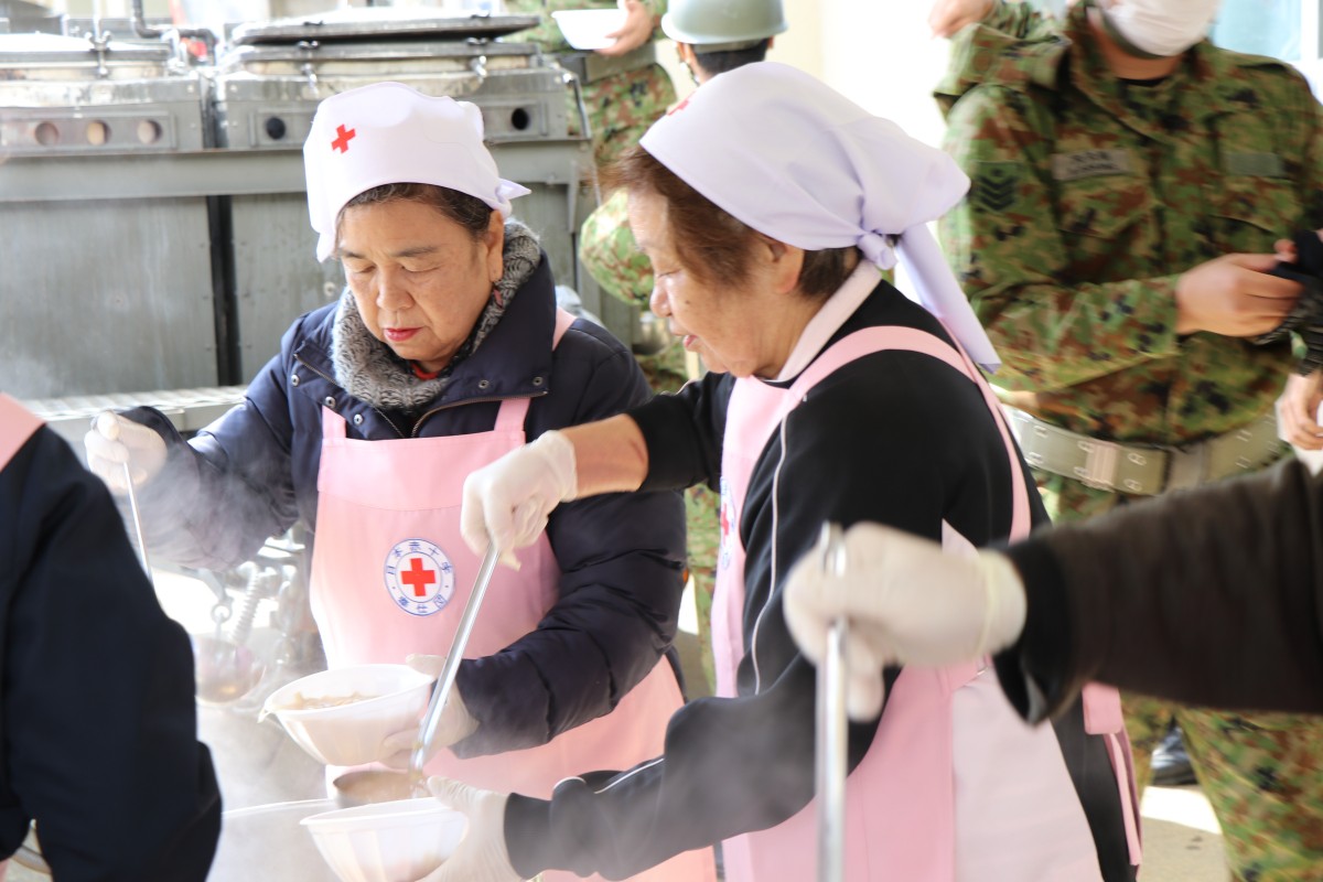 ピンク色の生地に赤十字マークが入ったエプロンを着た女性たちが紙皿に料理をよそっている写真