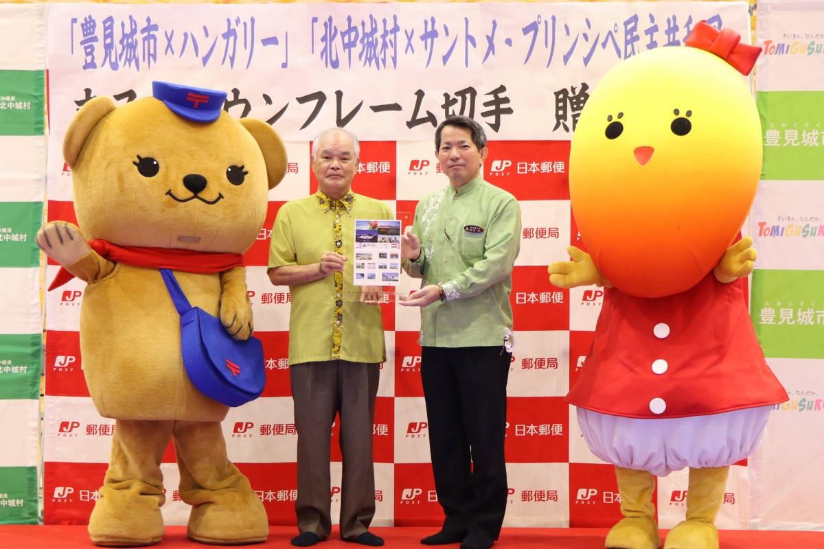郵便配達員の恰好をした熊の着ぐるみと黄色の顔をした赤い服を身を着ている着ぐるみの間で二人で紙を持っている男性の写真