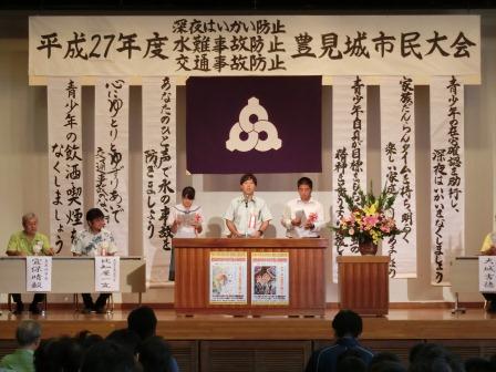 豊見城市市民大会と看板が掲げられているステージの上で制服を着た男女3人が立ってスピーチをしている写真