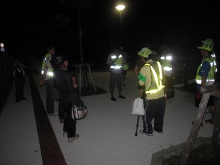 夜中の屋外の広場で反射板をついた安全服を着た人達が輪になって会話をしている写真