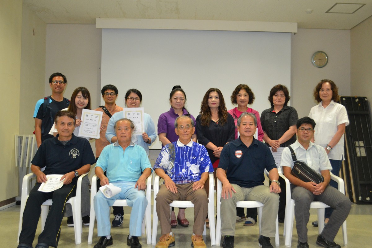 屋内で男性5人が椅子に座りその後ろに9人の男女が立ち修了証を持っている写真