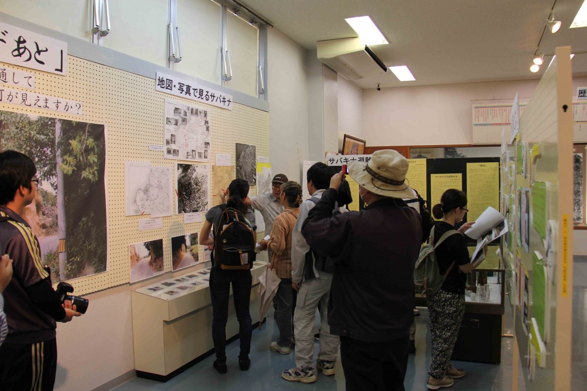 地図や写真の紙が貼られた展示室に、参加者たちが集まって説明を聞いている写真