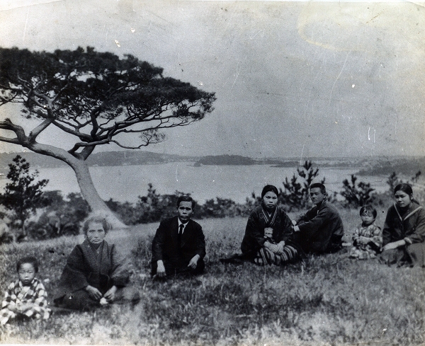 松の木と湖を背に、様々な服装の人たちが座ってくつろいでいる白黒写真