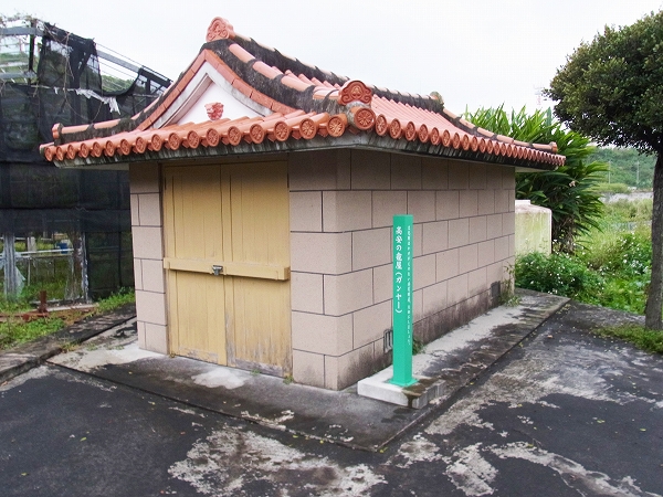 横に緑色の案内の柱が立てられている、瓦屋根と閂がある扉がついた小屋の写真