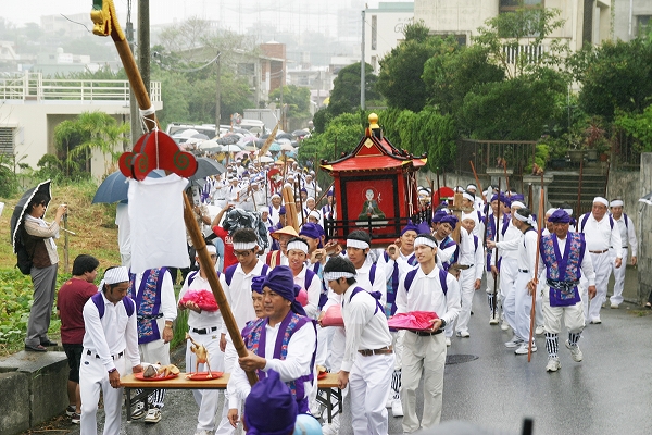 白い衣装に紫色の帽子やたすきを身に着けた人たちが、住宅街を練り歩いている写真