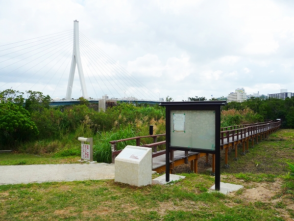 遠くに大きな橋が見え、手前に掲示板と木の遊歩道と説明版が置かれている写真