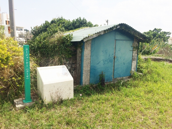 「コージャーヤー」と書かれた案内表示と白い説明文の横に、青い扉がついたトタン屋根の小屋がある写真