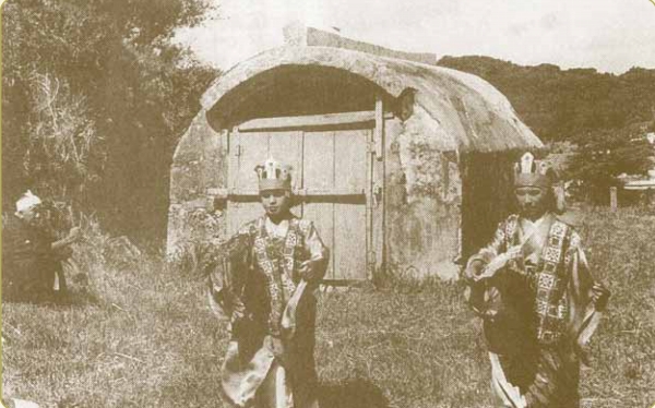 石でできた丸い屋根がついた小屋の前に、民族衣装を身に着けた二人の人が立っている写真