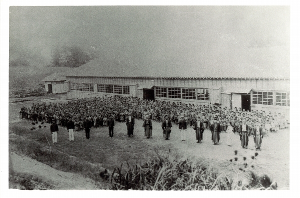 大きな平屋の建物の横に、何人もの人が整列して並んでいる白黒写真