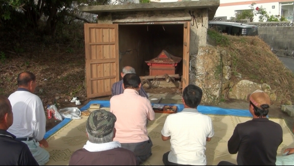 開いた小屋の中にある赤い祠にむかって、参加者たちが座って祈りをささげている写真