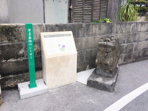 字名嘉地のシーサーと書かれた緑の柱と、隣に説明文がある台座があり、その隣に口を大きくあけたシーサーの石像が置かれている写真