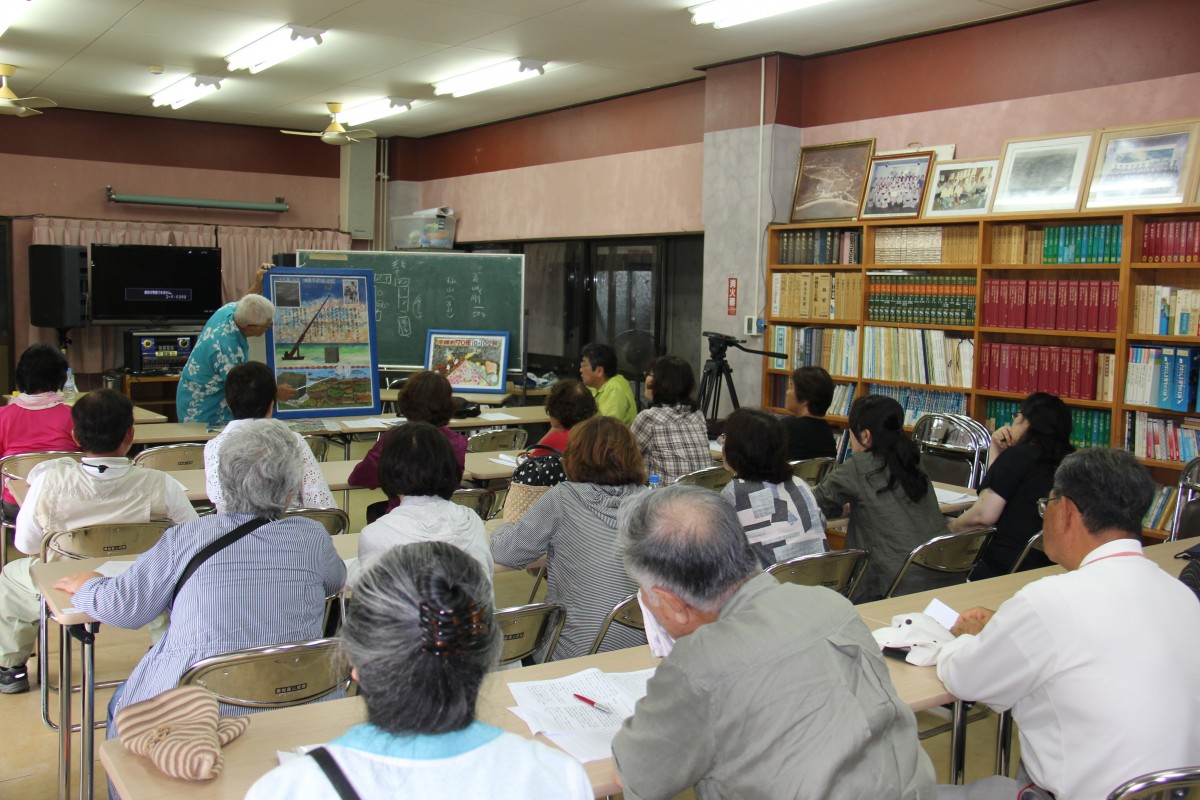 会議室の前方で、黒板や写真を見せながら話している人と、長机に座って説明を聞いている参加者たちの写真