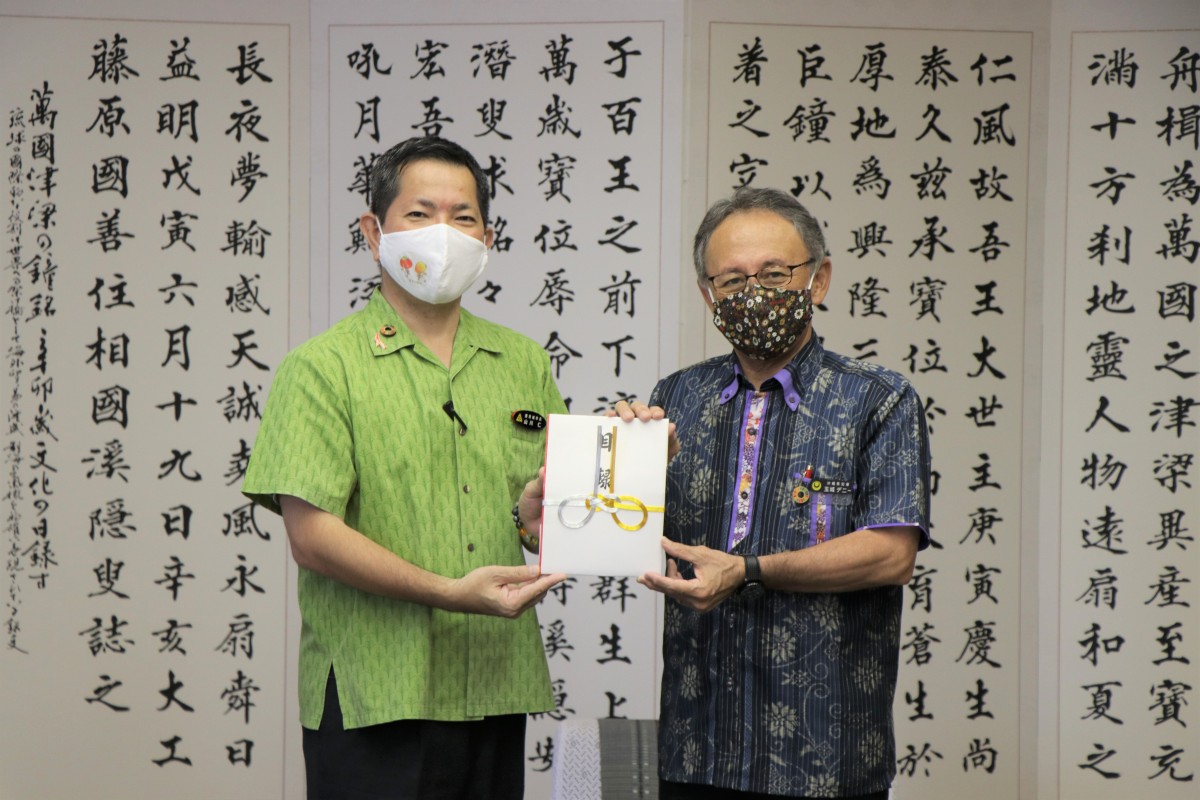漢字の紙が飾られている壁の前で、おじいさんと市長が封筒を持って並んでいる写真