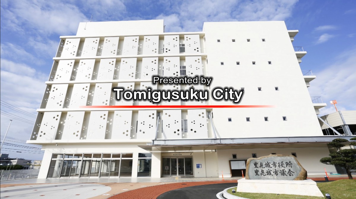 白い外壁が特徴的な豊見城市役所の外観と、その上に「Presented by Tomigusuku City」の文字が入っている写真