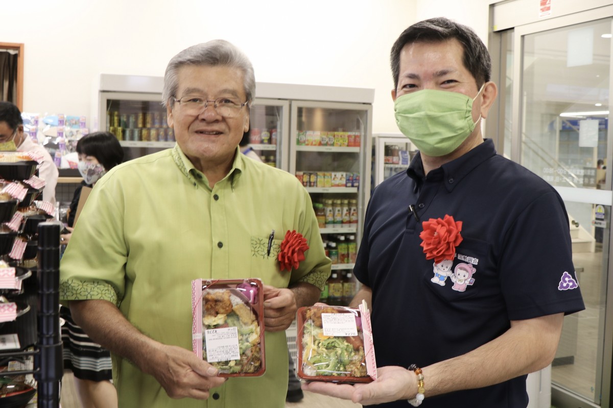 胸に赤い花の記章をつけ、商品のお弁当を手に取って見せながら微笑む男性二人の写真
