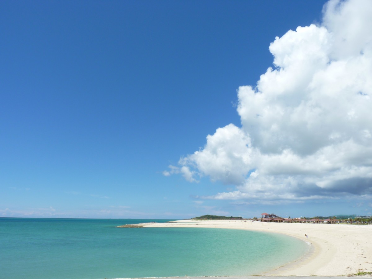 広い青空と大きな雲、弓形に広がる白い砂浜と青い海の写真
