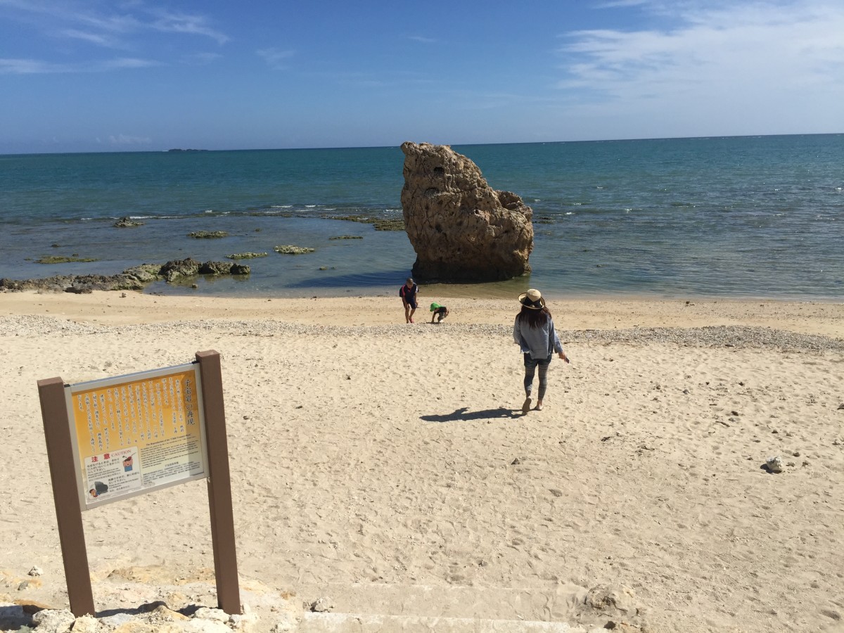 ビーチ入り口の看板と海に浮かぶ大きな岩と砂浜を歩く三人の写真